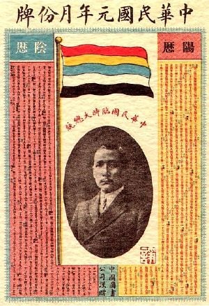 A calendar with a picture of a Chinese man in the middle. On top of it stands a flag with five horizontal stripes (red, yellow, blue, white, and black).