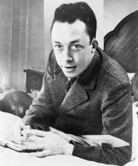 Albert Camus, gagnant de prix Nobel, portrait en buste, posé au bureau, faisant face à gauche, cigarette de tabagisme.jpg