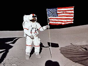 An astronaut in an Apollo space suit with red stripes on the arms and legs and down the helmet stands amid gray dust, grasping the pole of an American flag