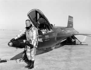 A black-and-white photo of Armstrong, with very short hair. He is smiling and is wearing a pressure suit and tall lace-up boots. Under his left arm he holds a bulky pressure helmet. He has black gloves on, and his right-hand rests on the nose of a dark-painted X-15 rocket plane with its canopy open. Armstrong and the plane are standing on a desert crust, and the plane's skids have left tracks in it.