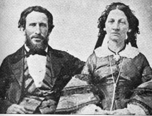 He has dark bushy hair and a beard and is wearing a three-piece suit with wide lapels and a bow tie. She has dark hair and wears a 19th-century dress with lace collar and bell sleeves.