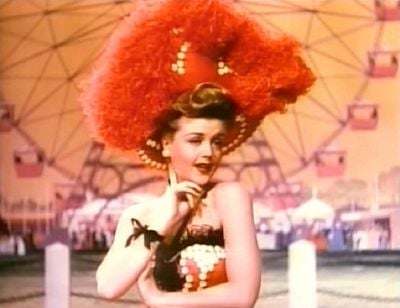 A young white woman, her arms exposed, wearing a large red feathered headdress. A fairground ride can be seen in the background.