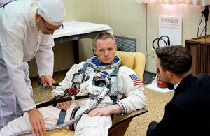 Armstrong, with short hair, partially reclining on a beige chair. He looks very serious. He is wearing a white space suit without a helmet or gloves. It has the U.S. flag on the left shoulder. Two hoses are attached. A technician dressed all in white is bending over him. A dark-haired, darkly dressed man has his back to us. He may be talking to Armstrong.