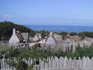 A modern-day photograph of a village consisting of small, primitive wooden houses. Most of the houses have thatched roofs. In the distance is a large expanse of ocean and a clear blue sky. The village is surrounded by a wall consisting of tall, thick wooden planks.