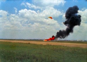 An indistinct photo of a smoke trail rising from an area of orange fire in a recently harvested field. A white and orange parachute is recovering a human figure above and to the right of the fire.