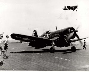 A Corsair on deck. A man stands nearby with fist upraised, giving a signal. There is another Corsair in the air above.
