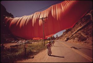 An enormous volume of fabric hangs from a wire across a valley. In the foreground is a telephone pole and several people looking up.