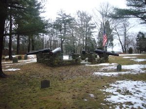 A monument featuring four black cannon barrels mounted on a stone wall in the middle of a small cemetery. The ground is partly covered with snow. Many trees stand in the background. The sky is cloudy.