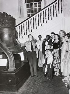 A man in uniform points to the Liberty Bell as a number of tourists, dressed in the fashions of sixty years ago, look on.