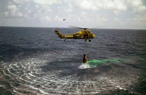 A green helicopter hovers low over the water, with the Mercury capsule suspended below. The helicopter has "Marines" written on it, and the number "44"