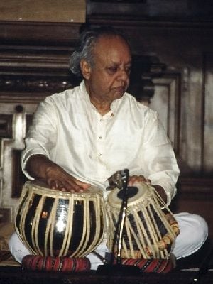 An old man sits cross-legged on the ground and rests his hands on two small drums.