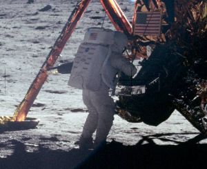 A grainy picture from behind of a human figure in white space suit and backpack standing in front of the Lunar Module on the surface of the Moon. A landing leg is visible and the U.S. flag on the descent stage.