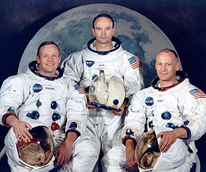 Three astronauts in white space suits. They are holding their helmets. All are light-skinned. Armstrong is smiling widely and wears his hair parted to the right. Collins has dark hair and looks the most serious. Aldrin's hair is very short. Behind them is a large photo of the Moon.