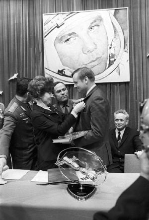 A black-and-white image. Armstrong has his left side facing us. He is holding a book and wearing civilian formal dress. A woman with bouffant hair is pinning a badge to his lapel. Two men in Soviet uniform and one in civilian garb are watching. On the wall in the background is a large photo of a cosmonaut. In the foreground on a table is a model of two spacecraft docking.