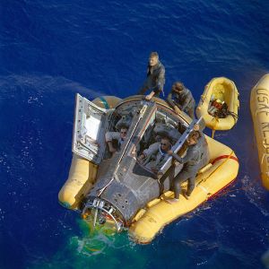 A dark gray Gemini capsule floats horizontally in blue water. It is supported by a yellow flotation collar. The hatches are open and the astronauts are visible sitting in their places wearing sunglasses. They are being assisted by three recovery crew in dark gray wetsuits.