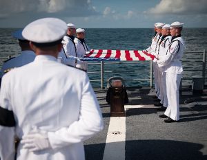 A squad of eight U.S. Navy personnel dressed in all-white uniforms hold a U.S. flag over a casket on the deck of a ship. The casket is carried on a dark wood plinth with several gold-colored badges. Much of the foreground is obscured by a senior officer with his back to us. Beyond is the sea.
