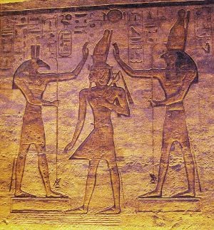Relief of a man with an elaborate crown between two figures who gesture toward the crown. The figure on the left has the head of an animal with square ears and a long nose, while the one on the right has a falcon's head.